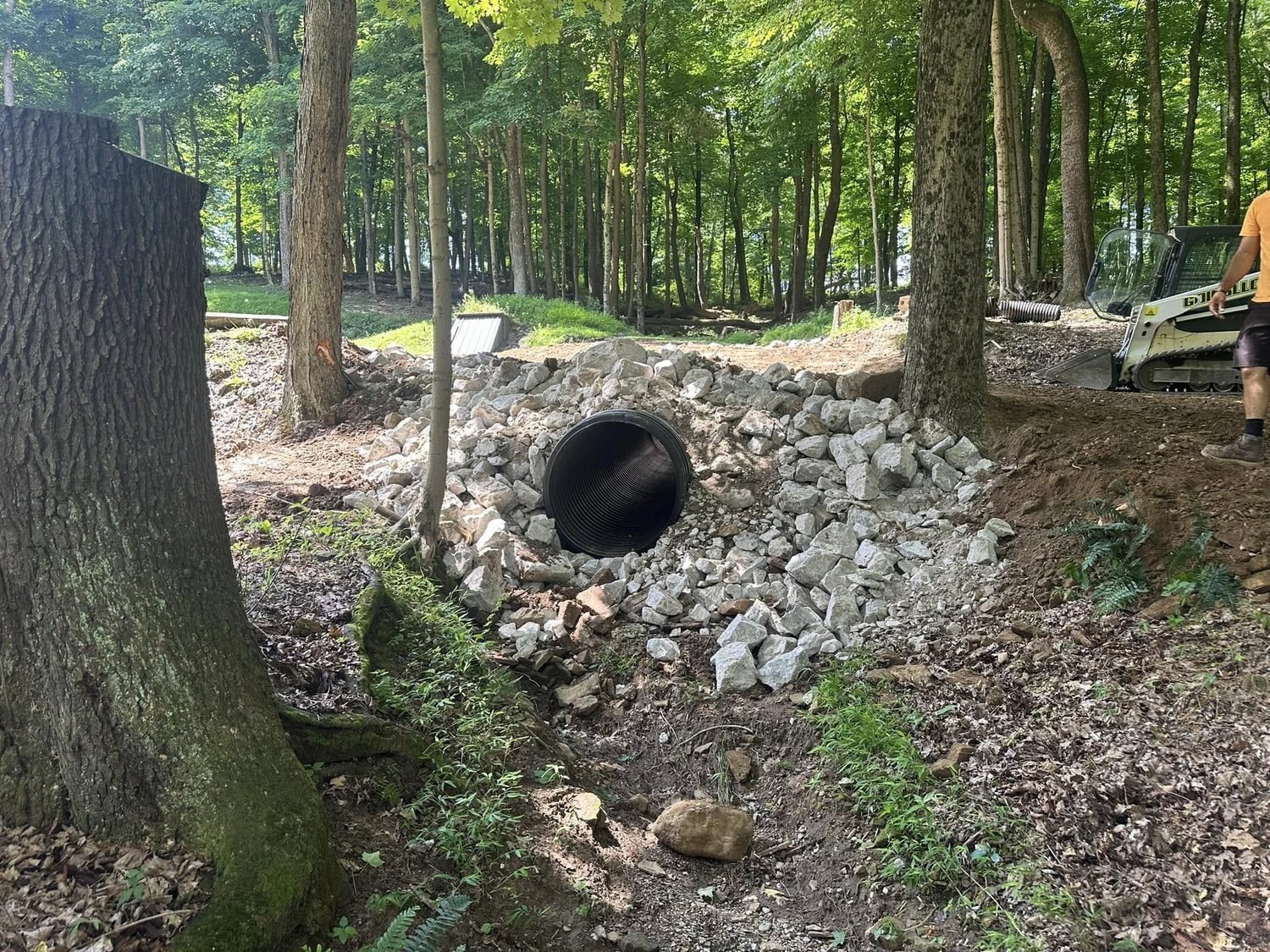 New culvert outlet set in a bed of rip-rap stone in the woods with a skid steer working alongside it