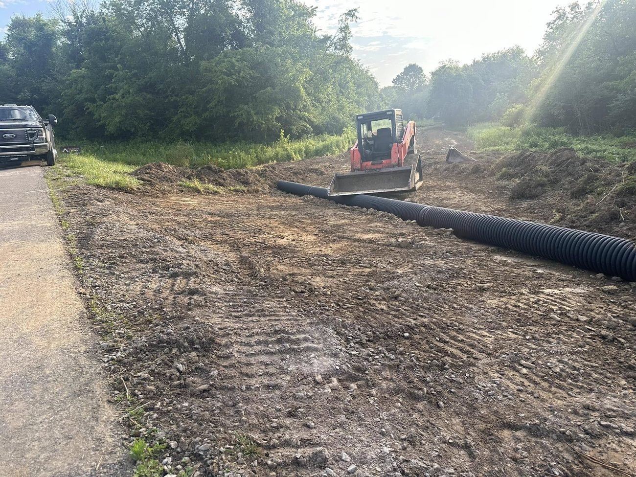 Long black corrugated culvert pipe staged along a roadside with a skid steer and truck at the edge of the job