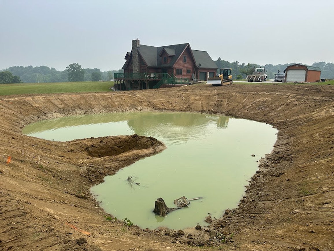 Newly dug farm pond with shaped banks filling with water in front of a log home
